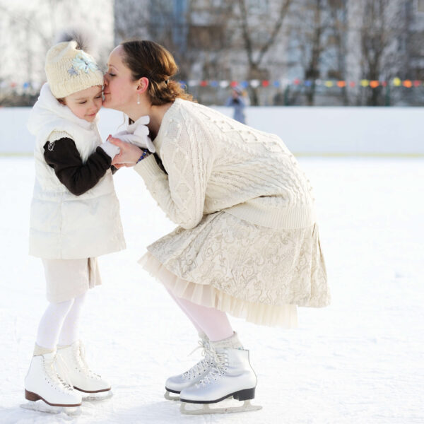 Eine Mutter k&uuml;sst liebevoll ihre Tochter auf einer Eisbahn. Winterkleidung und Schlittschuhe betonen die Szene. Climate Energy.