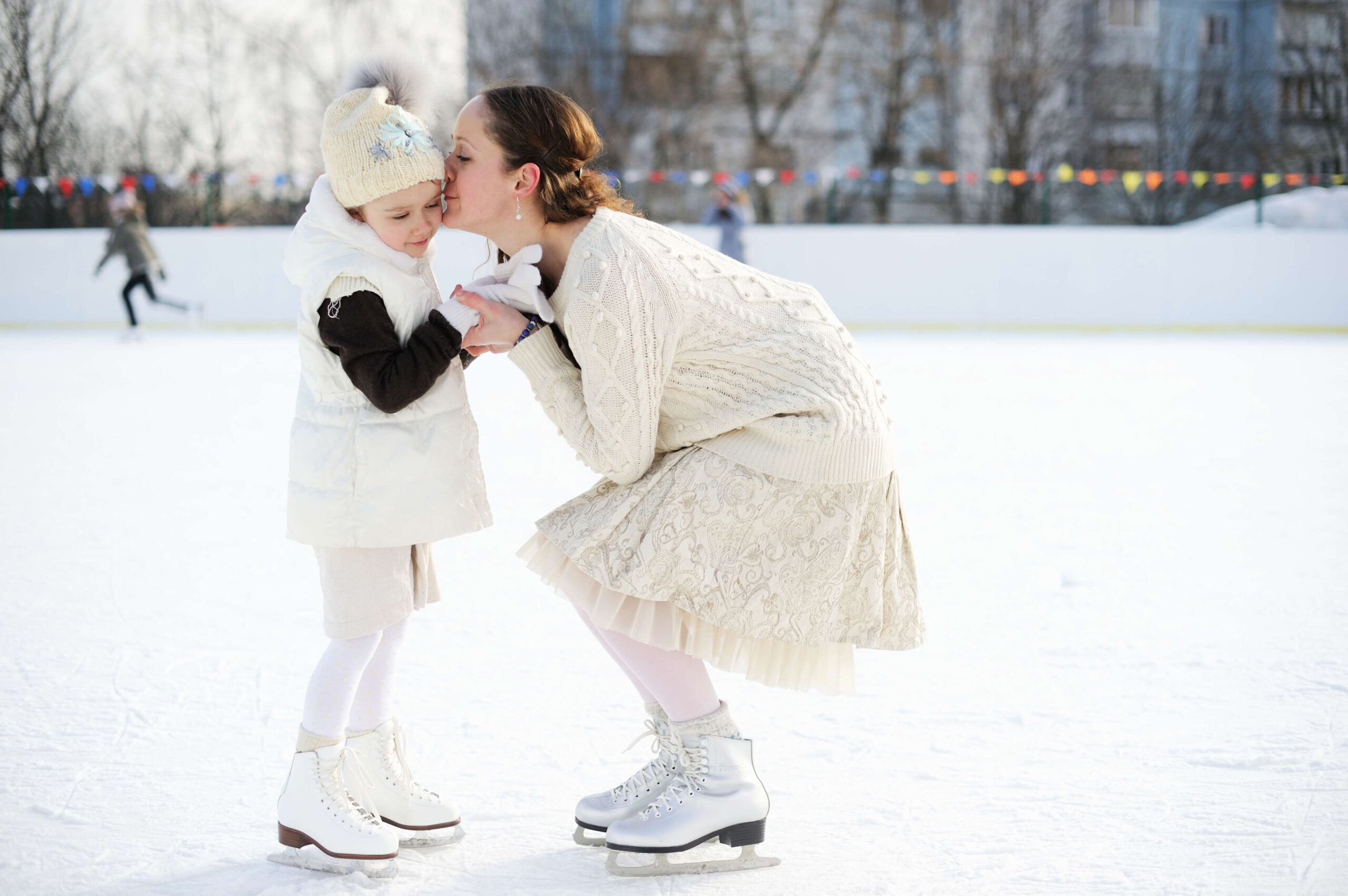 Eine Mutter k&uuml;sst liebevoll ihre Tochter auf einer Eisbahn. Winterkleidung und Schlittschuhe betonen die Szene. Climate Energy.
